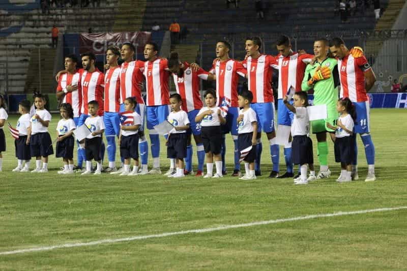 Tim sepak bola nasional Puerto Rico berdiri dengan tangan merangkul satu sama lain menyanyikan lagu kebangsaan.