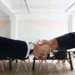 A handshake confirming a deal or acquisition occurring in a meeting room in a corporate office.