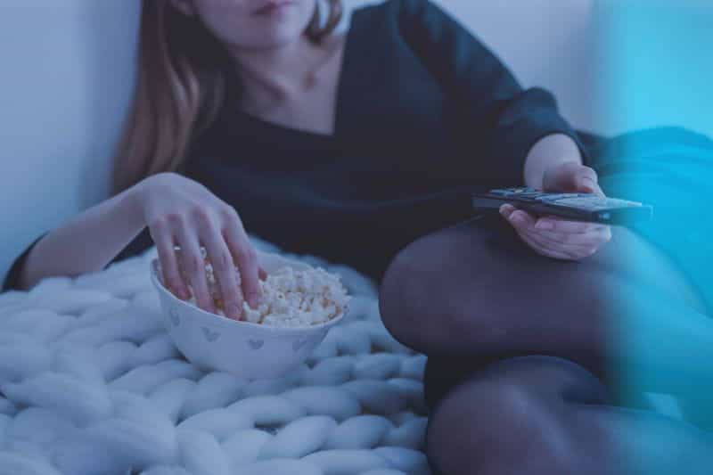A woman eating popcorn holding a remote control and watching TV.