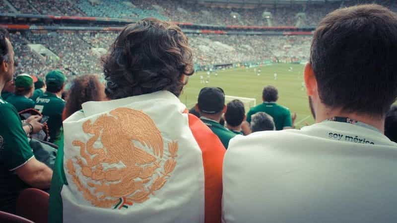 A soccer fan wrapped in the Mexican flag watches a live game in a crowded stadium.