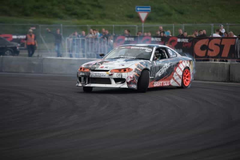 A racing car drifting on the track, kicking up smoke in front of an audience of spectators in the background.