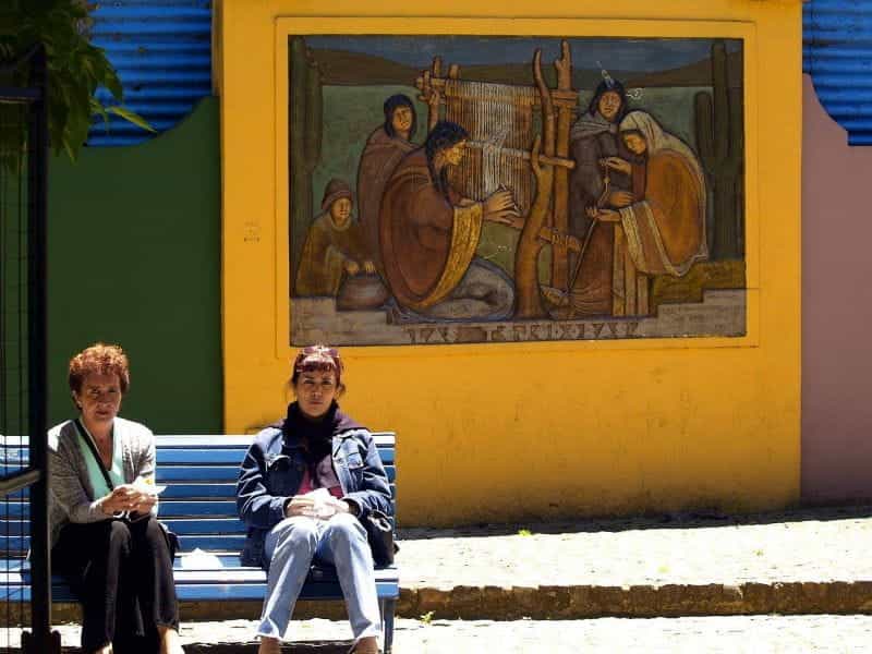 Two women sit on a bench in front of a mural in Buenos Aires, Argentina.