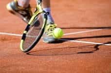 A tennis player dribbling a ball with their racket right next to their feet.