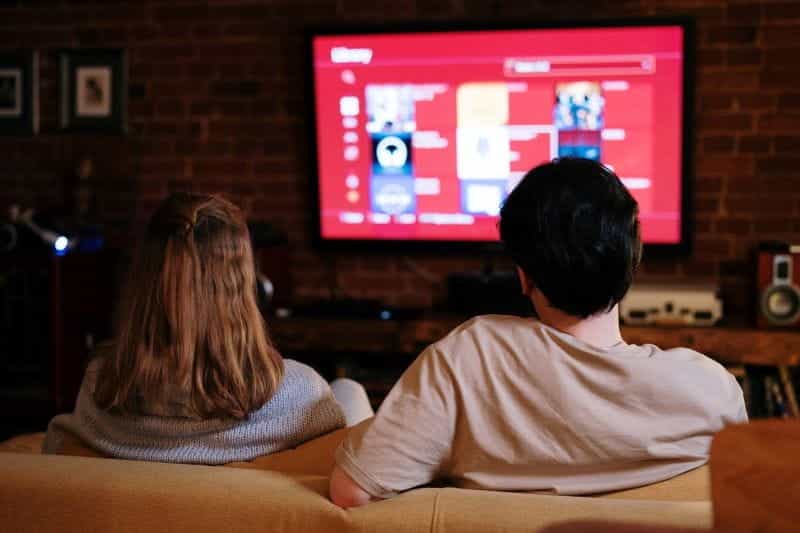Sepasang suami istri menonton TV, duduk di sofa di ruang tamu.
