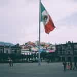 A Mexican national flag waves in a town square, in front of colorful buildings.