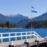 The Argentinian flag waves on a flagpole in front of a body of water and near a white fence.