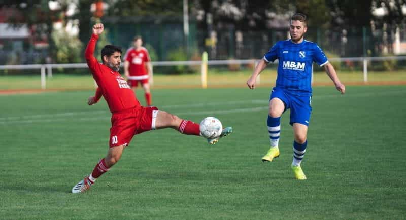 Two footballers playing on a pitch in red and blue kits.