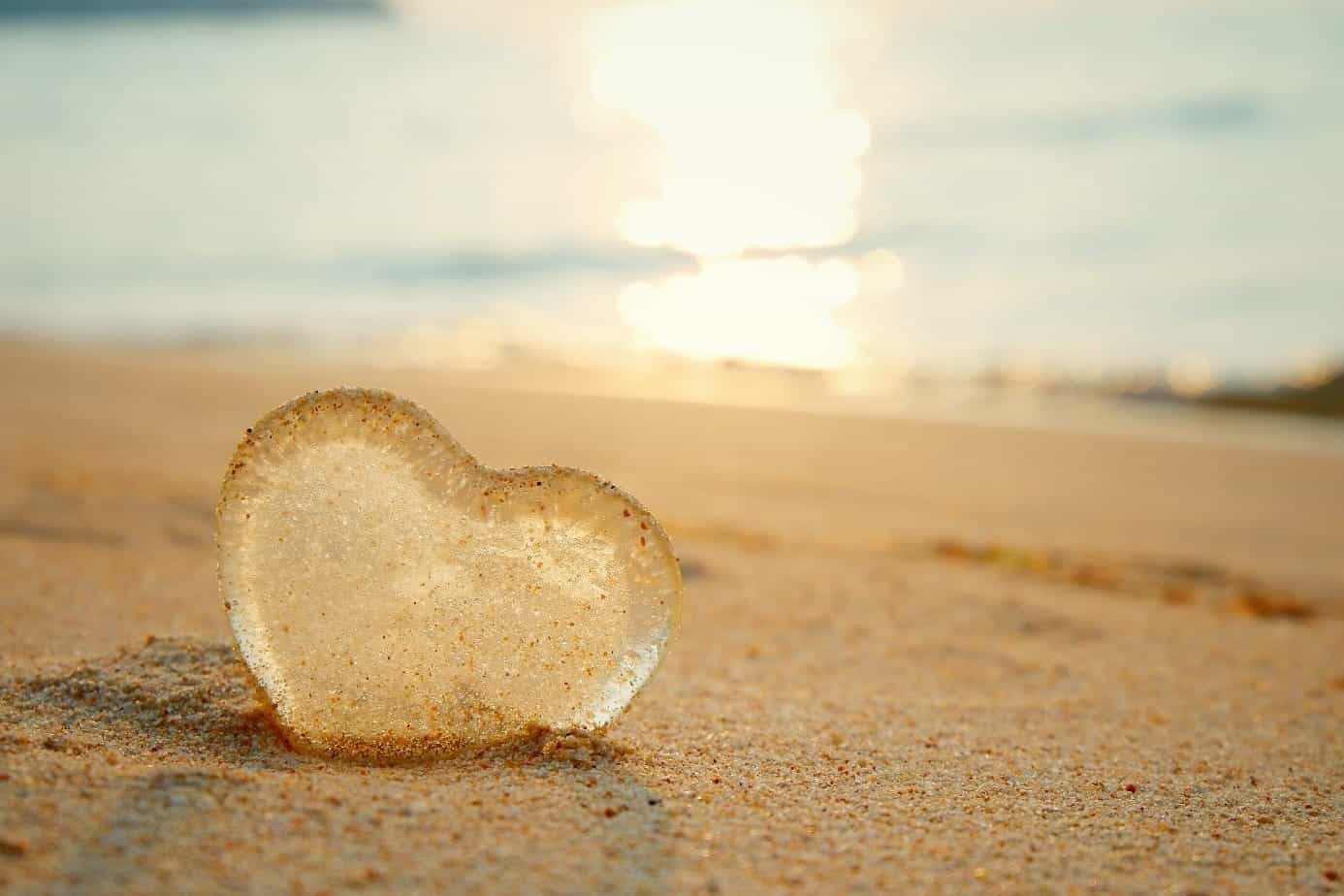 A glass heart sits alone on a sandy beach with the sun setting in the background