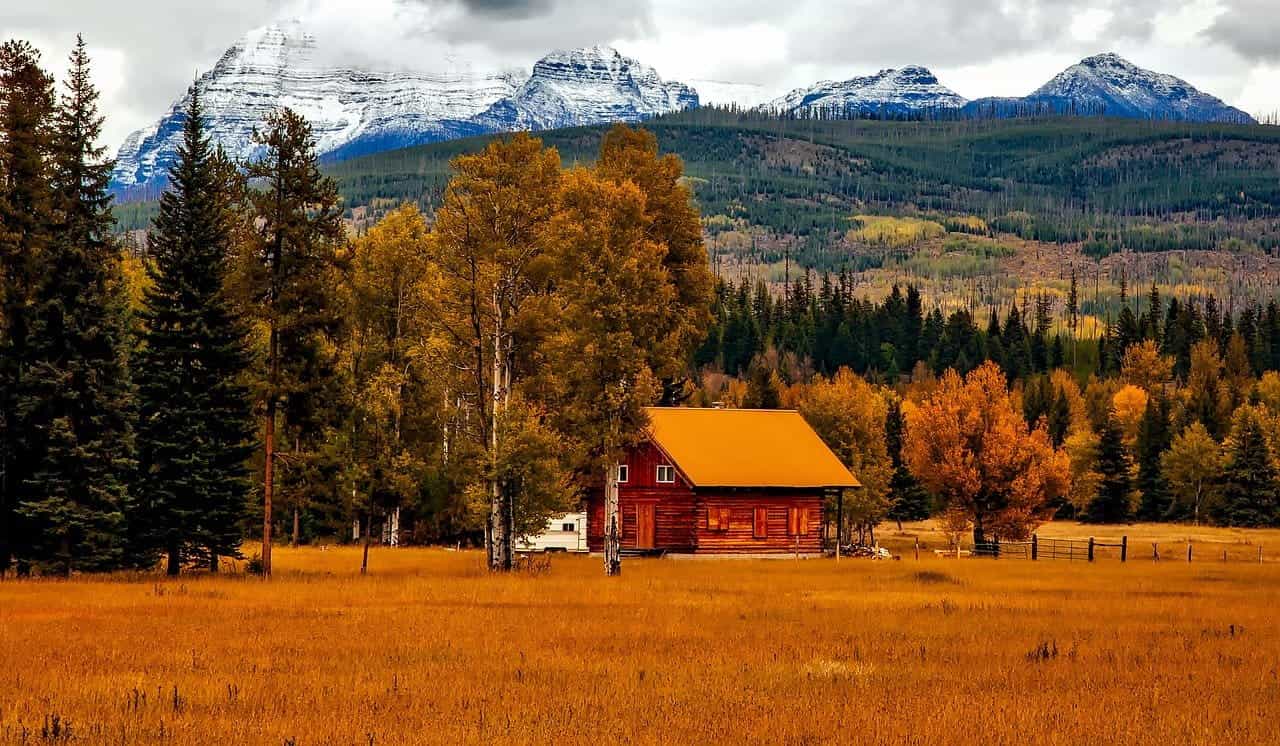 Rural Colorado in the fall, featuring a small red cabin surrounded by dense forests and towering, snow-capped mountains in the background.