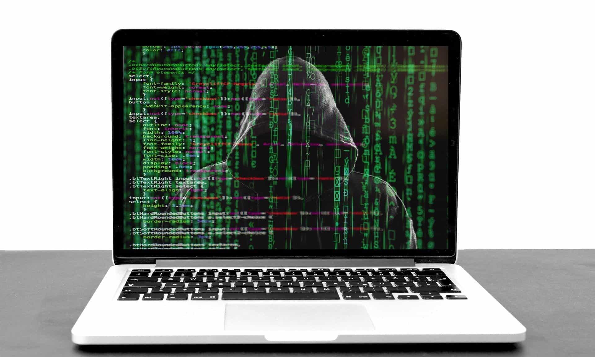 Open laptop computer with the screen covered in green hacker code and hooded character behind the code