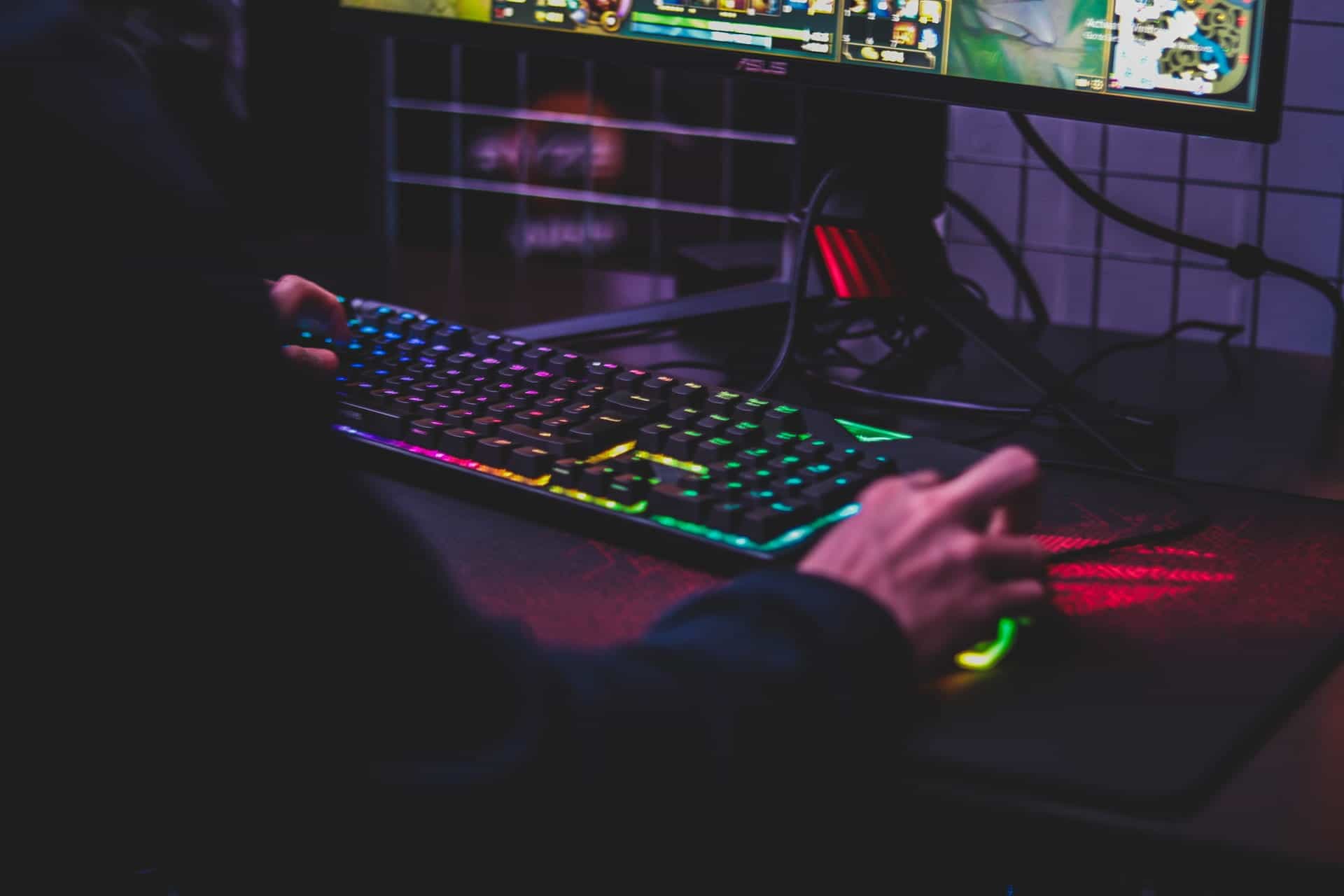 A closeup of a colorfully-lit gaming keyboard and hand as a person plays an immersive video game on a large computer monitor.