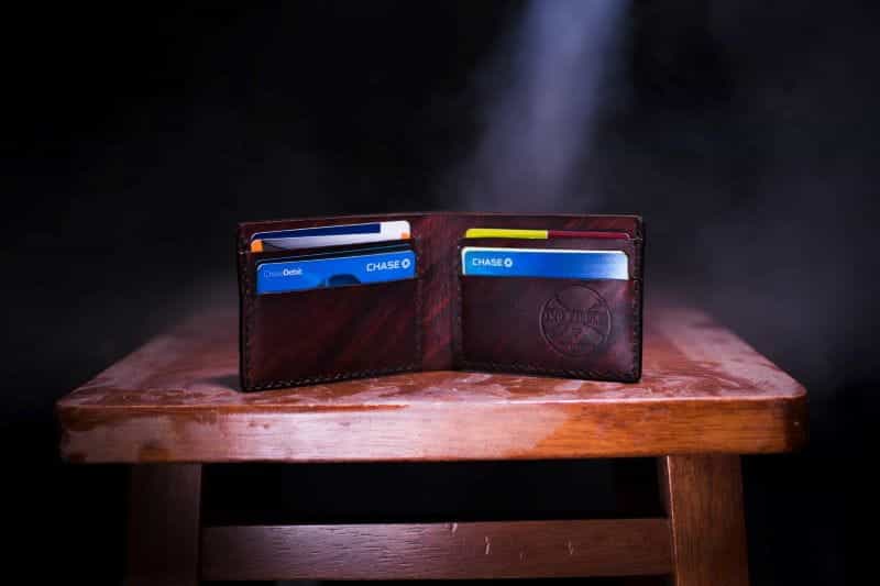 A brown, bifold leather wallet sits open on a wood table, revealing rows of payment cards inside.