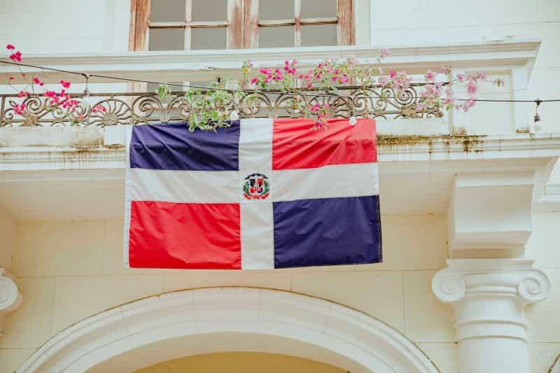 The Dominican Republic flag hangs on a balcony surrounded by flowers.