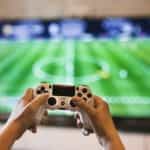 A video games controller is held up in front of a screen showing a football game.