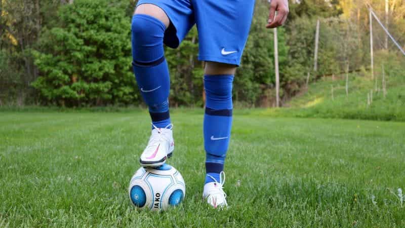 A person in a blue football kit with white boots with a football on a grass pitch.