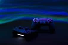 Two video game controllers resting near one another in front of a blue and green neon background.