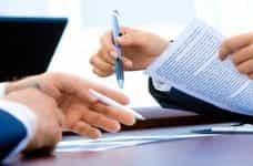 Two different hands each holding a pen as they review and sign an agreement on paper.
