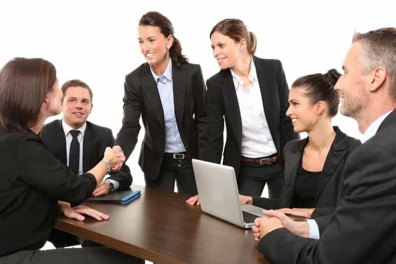 A group of people having a business meeting, with two women greeting each other and shaking hands.