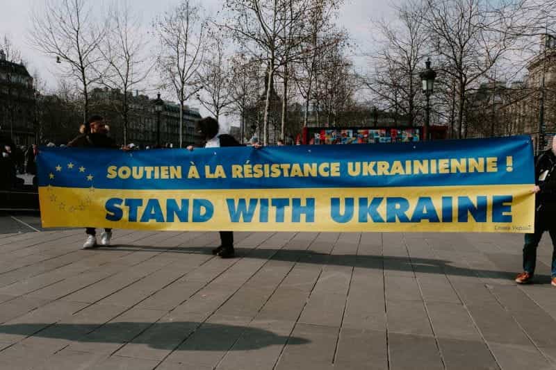 Banner blue at the top yellow at the bottom is held at either end. The copy in Ukrainian and English reads Stand With Ukraine.
