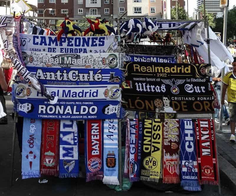 An open-air market stall with European club football merchandise including shirts, hats, and scarves.