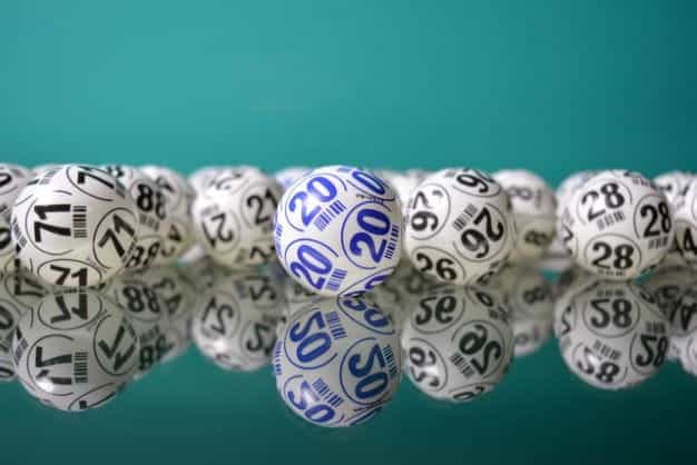 Lottery balls with black and blue numbers sit on a reflective surface.