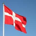 The Danish flag, white cross on red background, on a flagpole with blue sky.