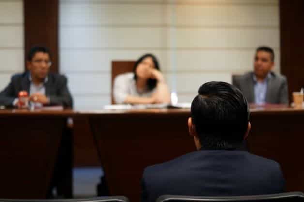 A group of people sit together in a council meeting as someone presents information.