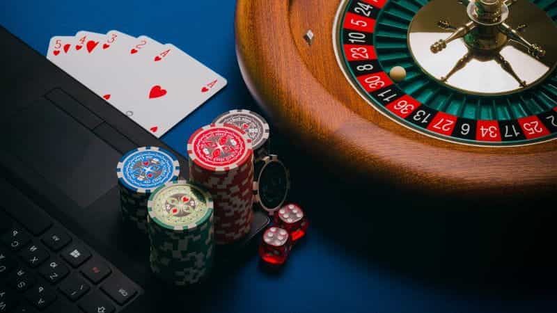 A roulette wheel, playing cards, poker chips and dice next to a laptop.