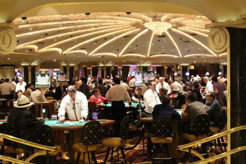 Inside a casino set out with games tables, croupiers in white jackets deal to seated players.