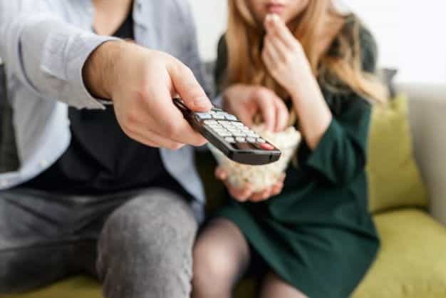 A man pointing a remote at a television, while a woman eats popcorn.