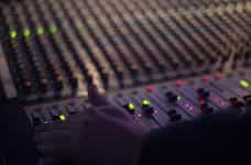 A wide, state of the art mixing console in a recording studio or music venue, with a sound engineer’s hand hovering over several faders.