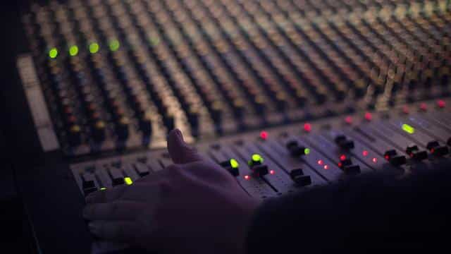 A wide, state of the art mixing console in a recording studio or music venue, with a sound engineer’s hand hovering over several faders.