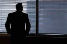 A man in a suit with his back facing the camera as he stares out of the blinds in a tall office building.