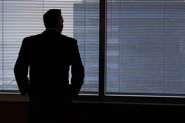 A man in a suit with his back facing the camera as he stares out of the blinds in a tall office building.
