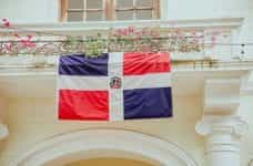 The Dominican Republic flag hangs outside an ornate building surrounded by vines and flowers.