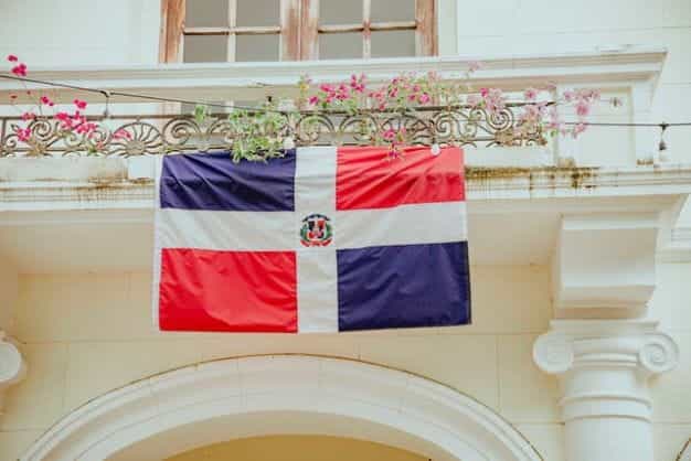 The Dominican Republic flag hangs outside an ornate building surrounded by vines and flowers.