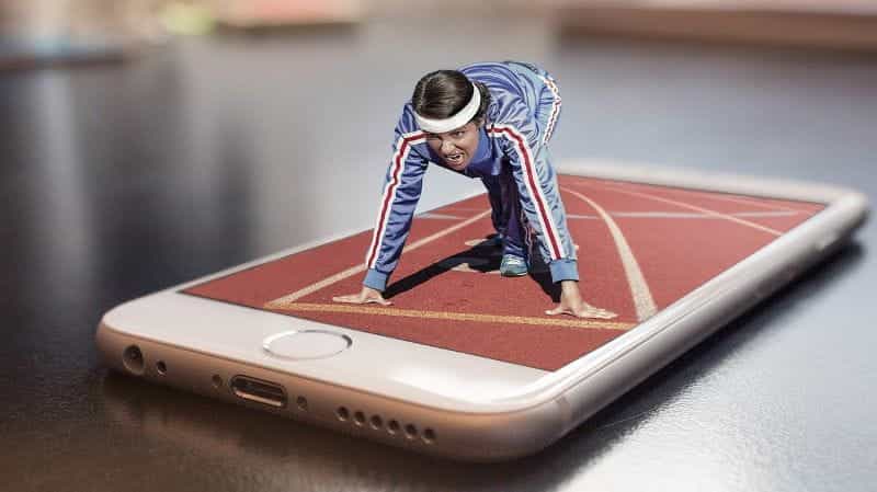 A smartphone lying flat on its back with the screen facing upwards, out of which a 3D runner athlete is emerging, poised at the ready to run a race.