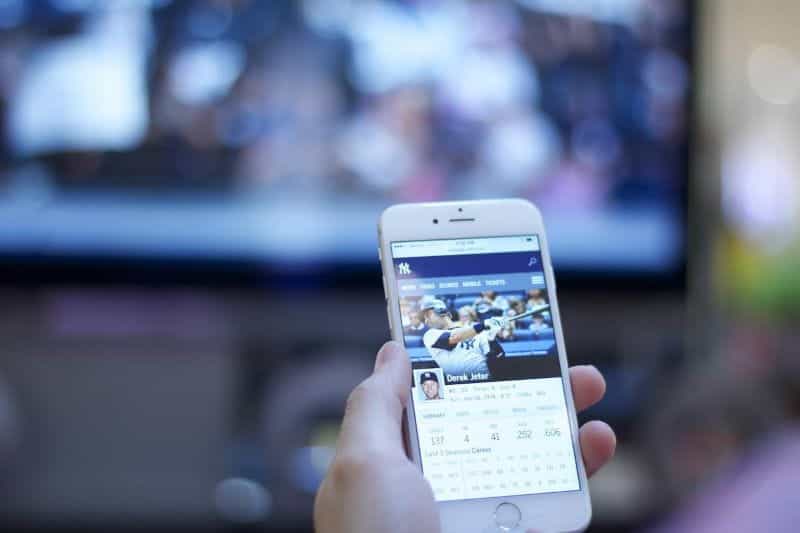 A person holding a smartphone displaying the stats of a baseball player on it in front of a television showing a sports match.