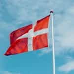 The Denmark flag hoisted on a pole against a blue sky backdrop.