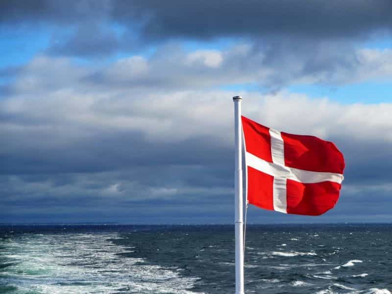 The Denmark flag hoisted on a pole against an ocean backdrop.