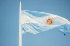 The blue and white Argentine flag waves against a blue sky, on a flagpole.