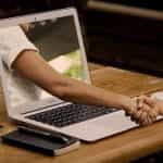 Someone shaking the hand of another person whose arm is coming out of the screen of a laptop, placed on a table and flanked by a smartphone, notebook and coffee mug.