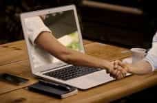 Someone shaking the hand of another person whose arm is coming out of the screen of a laptop, placed on a table and flanked by a smartphone, notebook and coffee mug.