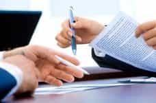 Two people holding pens and sorting through paperwork before signing a binding contractual agreement.