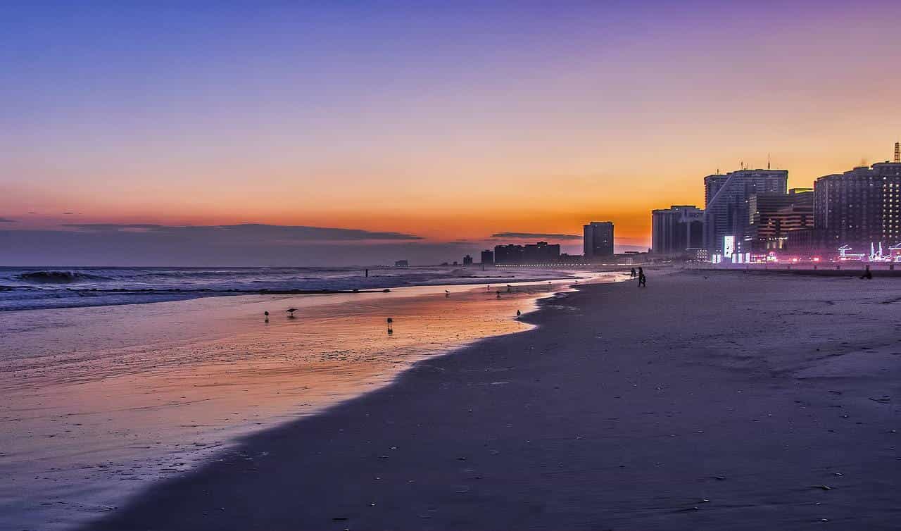 Pantai di Atlantic City, New Jersey saat matahari terbenam, dengan trotoar dan beberapa atraksi yang diterangi cahaya terlihat di sebelah kanan di kejauhan.