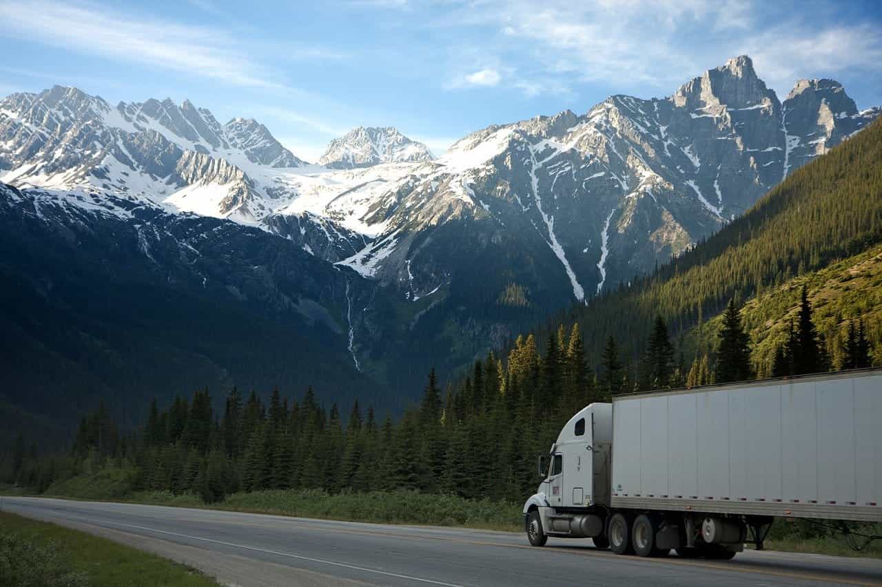 Sebuah truk barang besar melaju di jalan di pedesaan British Columbia, Kanada, dengan pegunungan tinggi dan megah terlihat di latar belakang.