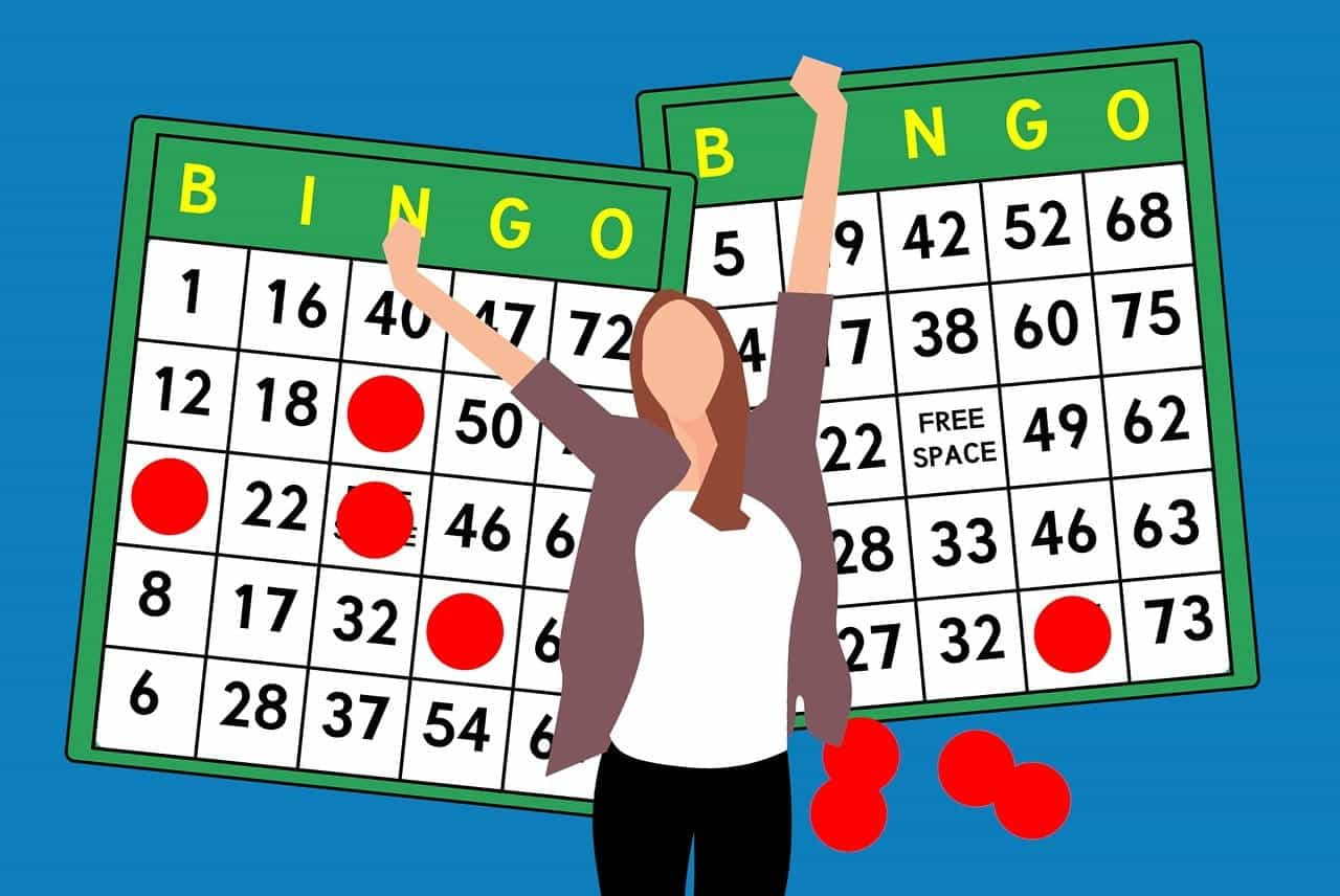 Seorang wanita mengangkat tangannya dalam perayaan sambil berdiri di depan dua kartu bingo, di mana spidol merah menunjukkan nomor bingo yang menang untuk permainan tersebut.