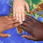 Three hands of people of different races coming together in unity above a map of the Earth.
