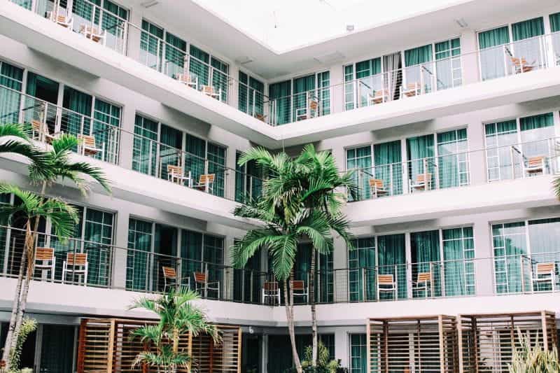 Fasad hotel dengan balkon dan kursi, serta pohon palem tropis.