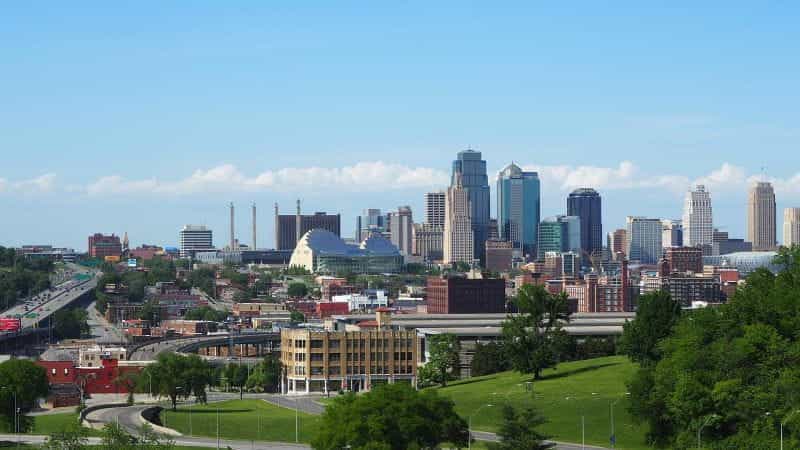 Cakrawala Kansas City, Missouri terlihat dari jarak dekat, menampilkan beberapa gedung tinggi dan pencakar langit yang diapit oleh jalan raya dan taman hijau.